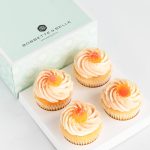 Four vanilla cupcakes topped with peach buttercream and fuzzy peach candies in a pastel bakery box.